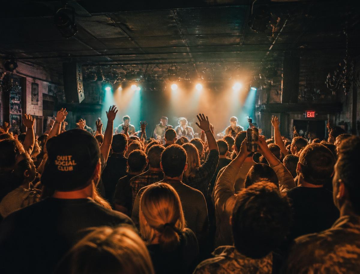 A packed intimate club audience with hands raised toward a lit stage